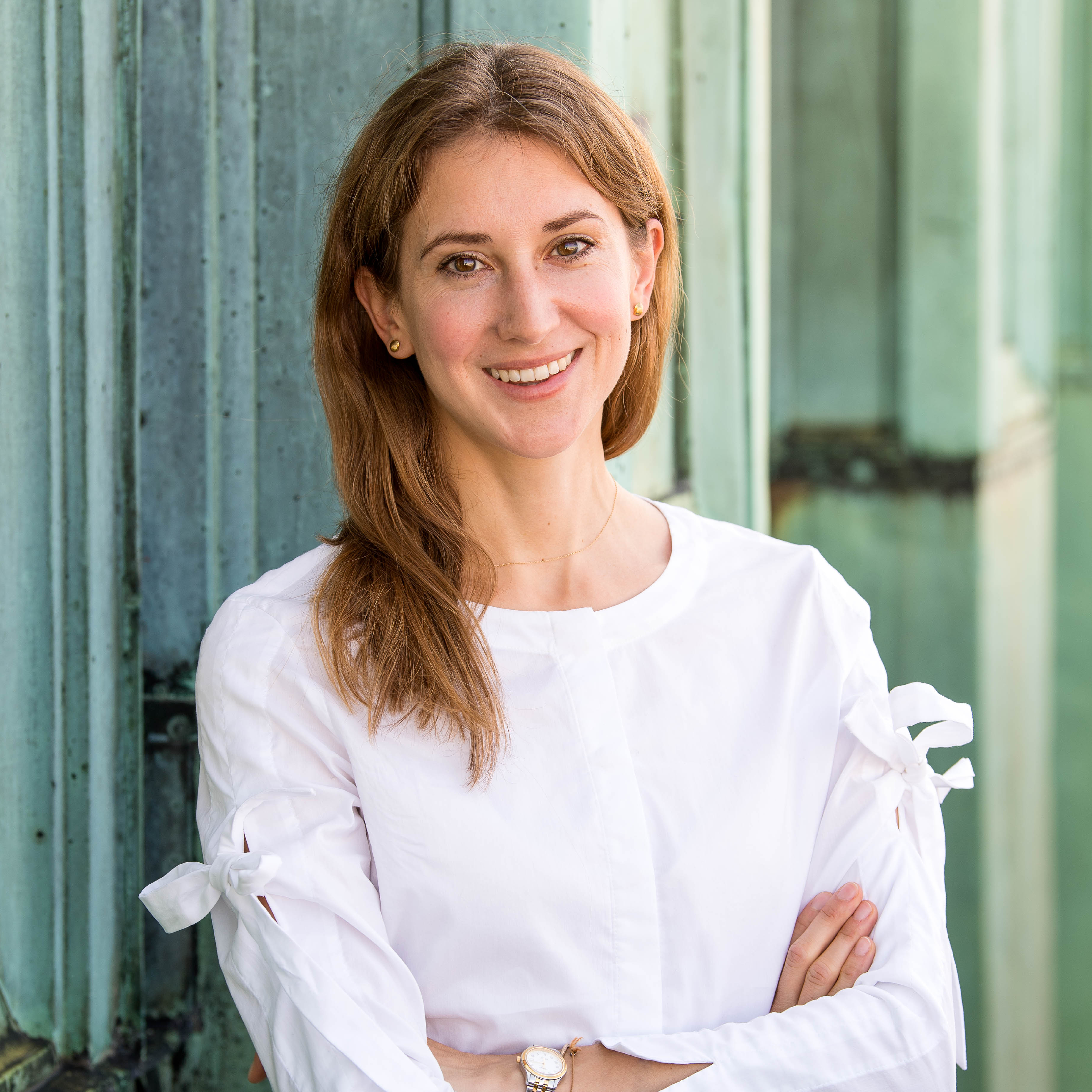 Lächelnde Frau in einer weißen Bluse, die mit verschränkten Armen vor einer grünen architektonischen Kulisse steht